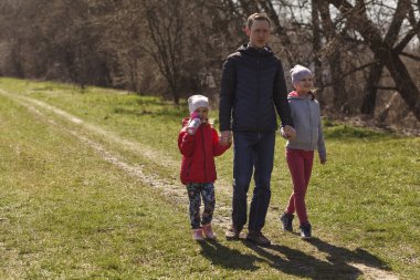 Doğada yürüyen iki kızı olan genç bir baba. İki çocuklu bir adam yolda yürüyor. Babalar çocukları ellerinden tutar..