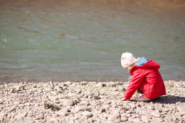 Nehirdeki küçük kız çakıl topluyor. Kırmızı ceketli bir çocuk nehir kenarında oturdu..