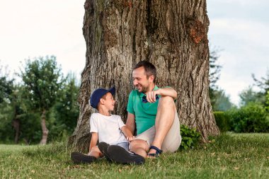 Gülümseyen baba ve oğul yaşlı bir ağacın altında çimlerin üzerinde oturuyorlar. İnsan ve çocuk doğada dinleniyor.
