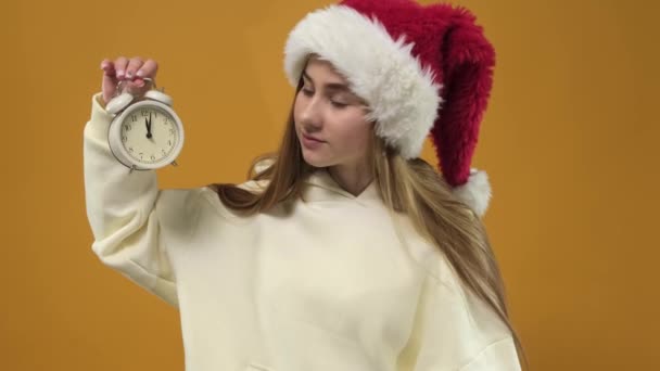 Μια νεαρή γυναίκα απομονωμένη σε ένα πορτοκαλί φόντο στούντιο κρατά ένα ξυπνητήρι και δείχνει το χρόνο. — Αρχείο Βίντεο