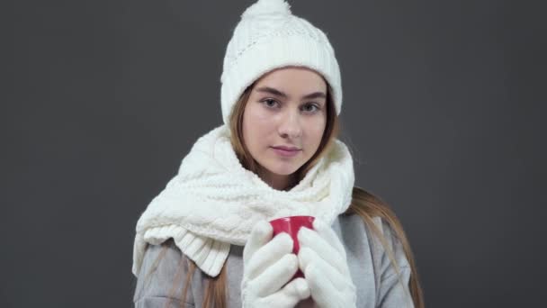 Portrait of a young beautiful girl in a white winter hat and gloves enjoying a delicious coffee while standing in the studio. — Stock Video