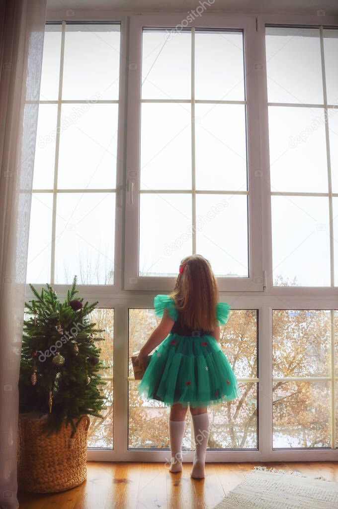 the little princess looks out the window. a girl in a green dress looks out of a large window. festive elegant fir tree with garlands. Merry Christmas and happy New Year. 