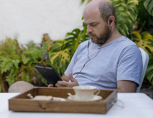 A man is staring intently at a digital device, listening headphones in his ears.The person may be participating in an online work meeting from his backyard or watching a video or getting information on his digital tablet. He has a serious attitude. D