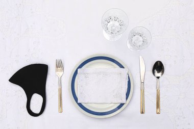 Üzerinde bir tabak, hassas bir peçete, bıçak, çatal, kaşık, su bardağı, şarap kadehi ve koruyucu bir koronavirüs maskesi olan beyaz masa örtüsüyle döşenmiş bir masa. Yukarıdan bakıldığında, insanların ve yiyeceklerin olmadığı bir restoran masası. Boşluğu kopyala.