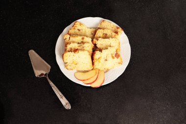 Üst Manzara. Elmalı turta, meyve dilimleri ve paslanmaz çelikten bir spatula içeren beyaz porselen tabağın yakın plan detayları. Hepsi siyah bir mermer masanın üzerinde..