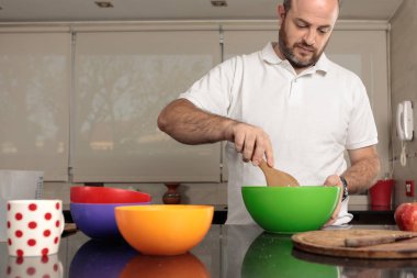 Beyaz bir adam, 30-39 yaşları arasında, karışımı bir kaseyle karıştırıyor, bir tahta kaşıkla, ev mutfağında elmalı turta hazırlıyor. Turtanın yapılışını gösteren birden fazla resim serisi. Dizinin bir parçası.