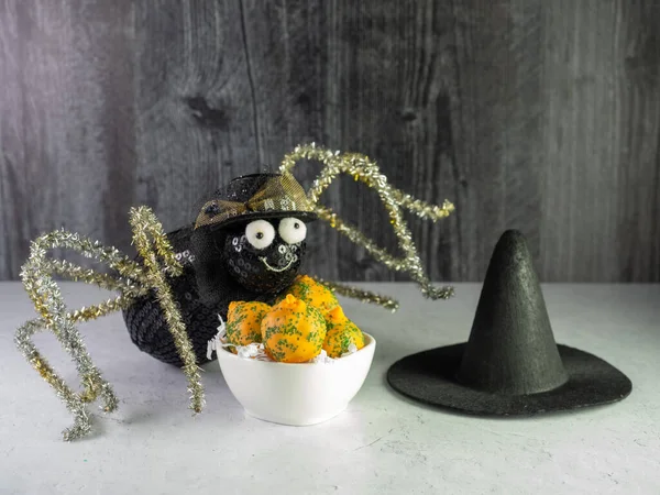Black Spider with gold pipe cleaner legs Posing with a white bowl full of orange and green Carrot Cake Balls and a black witch hat.  Trick or Treat for Halloween and fall!