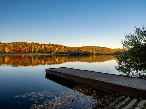 A boat dock in the foreground at Keystone Lake in West Moreland Country, nestled in the Laurel Highlands of Pennsylvania in the fall with the colorful trees reflecting in the lake and a nice blue sky.