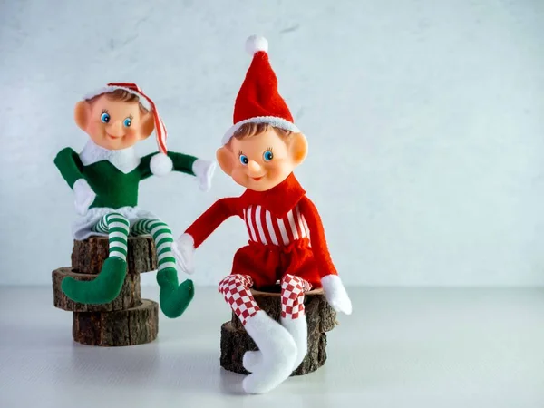 Two Christmas Elves sitting on wooden tree trunks pieces, one with a green outfit and one with a red outfit.  Holiday decor, Santa Hat, elf, red and green.