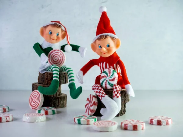İki Noel Elfi ve Kırmızı ve Yeşil Giysiler Ağaç üzerinde oturuyor, ellerinde daha fazla şekerle büyük şeker parçaları tutuyorlar. Tatil dekoru, Noel Baba şapkası ve nane şekeri..