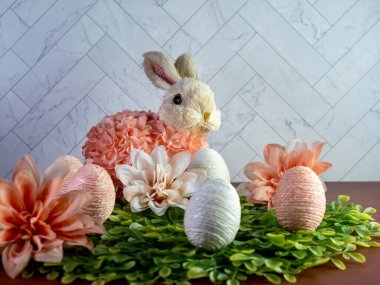 Yeşil yapraklardan oluşan bir yatakta oturan Paskalya tavşanı şeftali çiçekleri çiçek açıyor ve terra-cotta yüzeyinde bembeyaz iplikler ve şeftali rengi yumurtalar var..