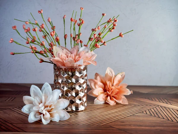 Peach dahlia flower blooms with a hammered metal vase filled with orange colored flower buds sitting on a striped wooden surface.  Simple, spring home decor.