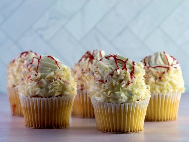 Üzerinde beyaz kremalı, yüksek kıvrımlı beyaz çikolatalı, üstünde kırmızı ahududu çiselemesi olan beyaz kapkekler, pembe mermer tezgahlı lezzetli tatlı ve ringa kemiği duvarlı..