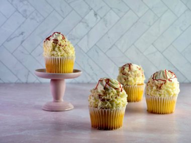 Üzerinde beyaz kremalı, yüksek kıvrımlı beyaz çikolatalı, üstünde kırmızı ahududu çiselemesi olan beyaz kapkekler, pembe mermer tezgahlı lezzetli tatlı ve ringa kemiği duvarlı..