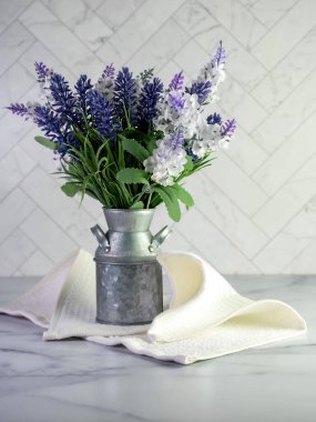 Basit bir bahar evi dekorasyonu için yapay mor lavanta ve leylaklarla dolu taşralı metal süt kutusu ve ringa kemiği fayanslı mermer tezgah..  