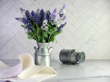 Basit bir bahar evi dekorasyonu için yapay mor lavanta ve leylaklarla dolu taşralı metal süt kutusu ve ringa kemiği fayanslı mermer tezgah..  