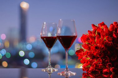 Yıldönümü veya Sevgililer Günü konsepti için renkli şehir ışıkları olan masada iki kadeh kırmızı şarap ve kırmızı güller..