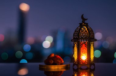 Üzerinde ay sembolü olan fener ve üzerinde gece gökyüzü ve şehir arkaplanı olan küçük hurma tabağı kutsal Ramazan ayı Kareem 'in Müslüman ziyafeti için ışık saçar..