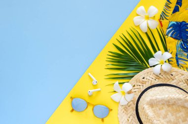Plaj şapkası, güneş gözlüğü, hindistan cevizi yaprakları, kablosuz kulaklık, fransizca çiçekleri ve sarı ve mavi arkaplanda renkli gömlekli yaz konsepti..