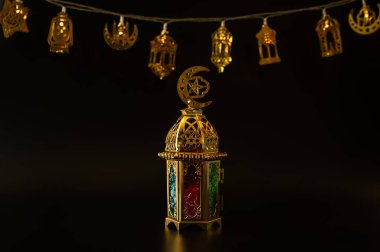 Renkli cam panellerle süslü bir altın Ramazan feneri Hilal ve yıldız ışıklarının önünde duruyor..