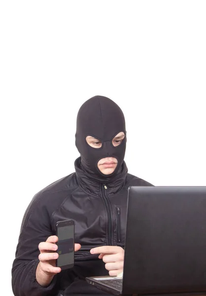 Siyah maskeli, cep telefonu ve dizüstü bilgisayarı olan bir adam internet dolandırıcılığıyla meşgul.