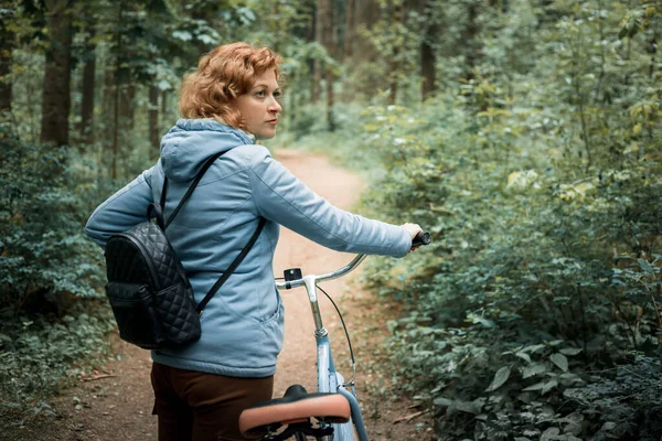 Une femme blonde vêtue d'une veste bleue marche dans la forêt avec un vélo bleu — Photo
