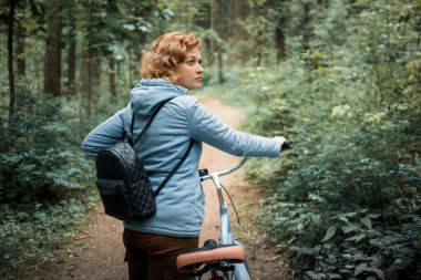Mavi ceketli sarışın bir kadın mavi bir bisikletle ormanda yürüyor.