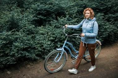 Mavi ceketli sarışın bir kadın mavi bir bisikletle ormanda yürüyor.