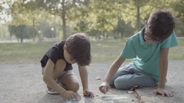 Δύο χαριτωμένα αγόρια κάθονται στο έδαφος και ζωγραφίζουν με κιμωλίες — Αρχείο Βίντεο
