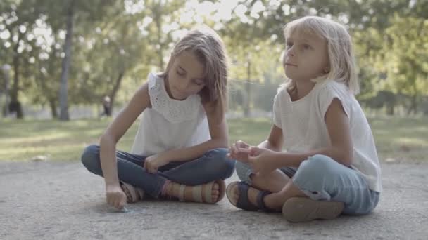 Twee blanke meisjes zitten gekruiste benen en tekening met krijt — Stockvideo