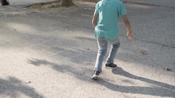 Menino de camisa azul, jeans e óculos jogando hopscotch — Vídeo de Stock