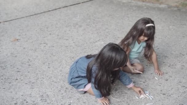 İki Latin kız kardeş asfaltta oturuyor ve tebeşirle çizim yapıyorlar. — Stok video
