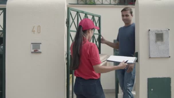 Női futár piros sapkában szállít csomagot az ügyfelek ajtajába — Stock videók