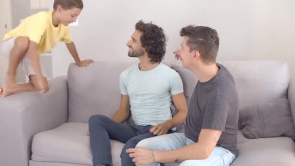 Wesoły chłopiec skaczący na kanapie do swoich dwóch ojców — Wideo stockowe