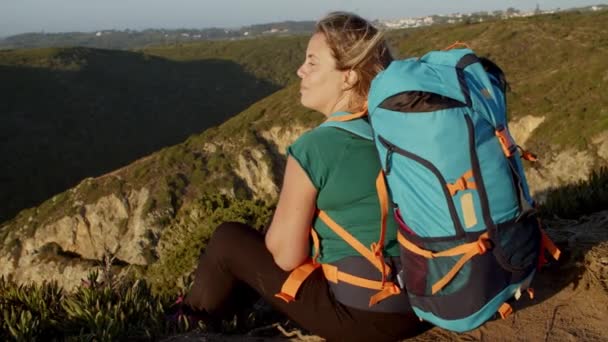 Lykkelig kvindelig backpacker sidder ved klippen, kigger på solnedgang – Stock-video
