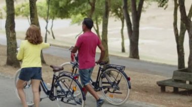 Parkta bisikletle yürüyen beyaz bir çiftin arka görüntüsü.