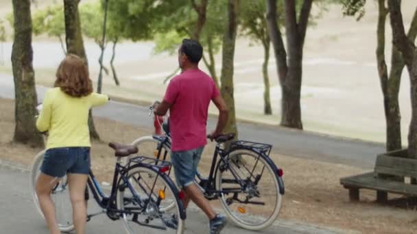 Vedere din spate a cuplului caucazian mers pe jos cu biciclete în parc — Videoclip de stoc