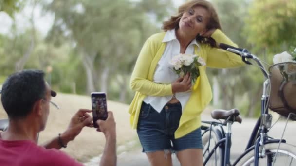 Вид спереди мужчины, фотографирующего жену с цветами в парке — стоковое видео
