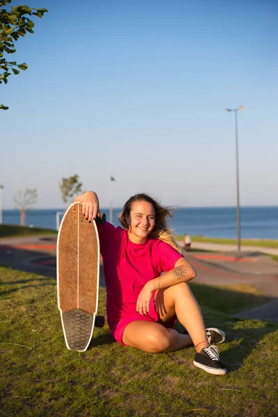 Portrait of smiling young woman with longboard sitting on grass — Stock Photo, Image