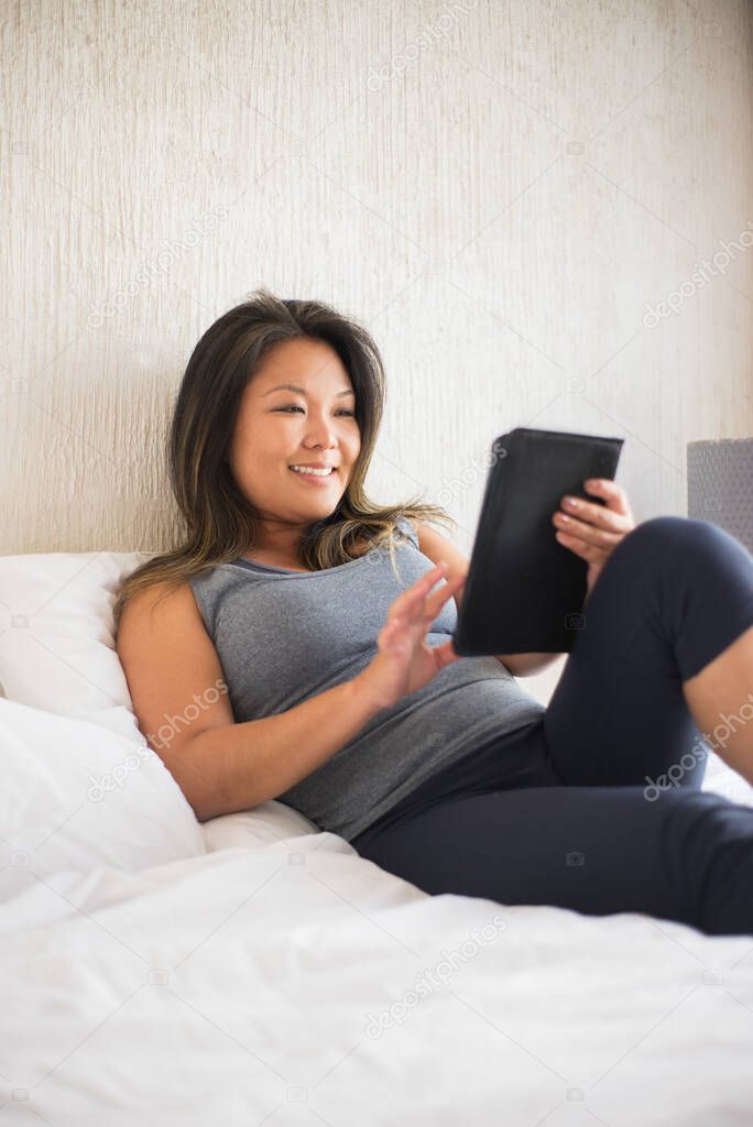 Smiling beautiful Japanese woman reading electronic book in bed