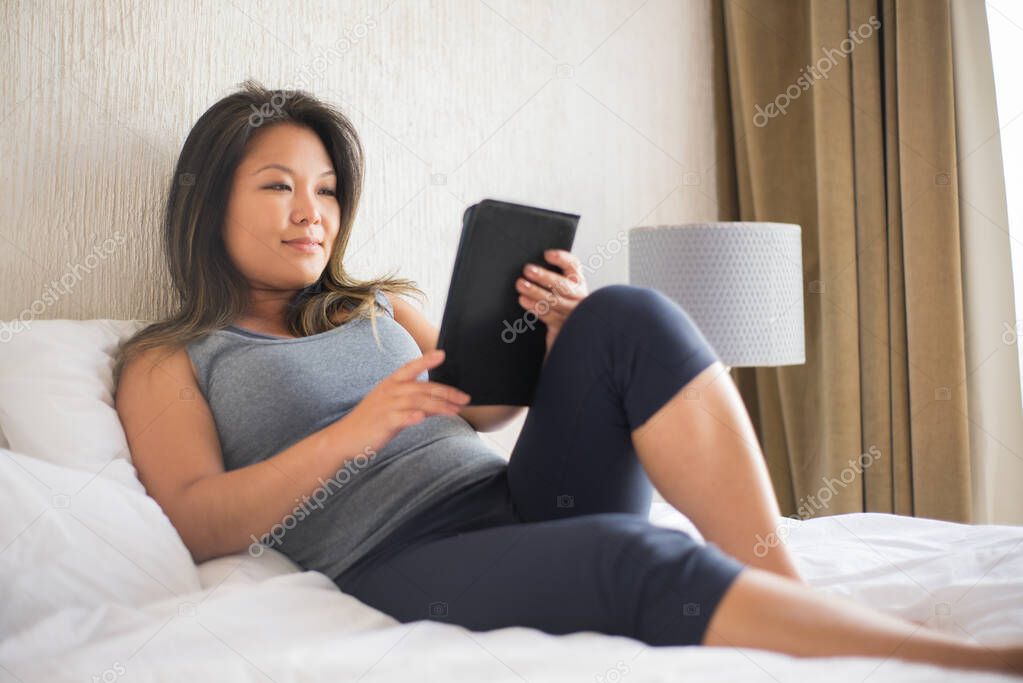 Calm Japanese woman reading electronic book in bed