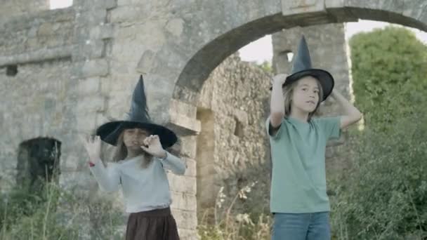 Niño y niña de pie en sombreros de bruja entre las paredes de piedra del castillo — Vídeo de stock