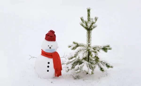 Sneeuwman met gezichtsmasker, winterhoed. sjaal op een witte achtergrond en staand bij een boom. — Stockfoto