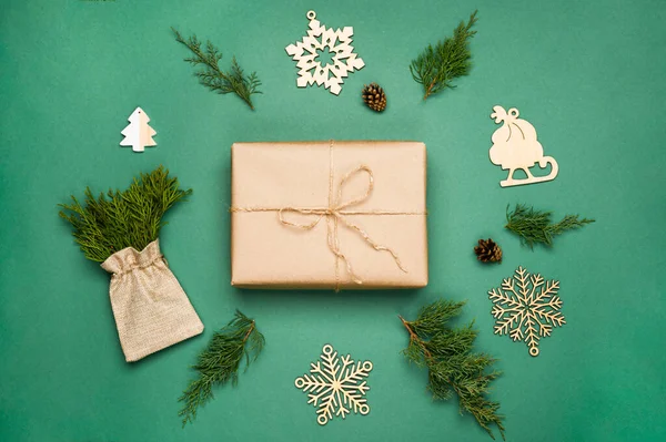 Skład z bukietem gałęzi iglastych w eko torbie, drewniane płatki śniegu, pudełko prezentów w papierze kraft, szyszki na zielonym tle. — Zdjęcie stockowe