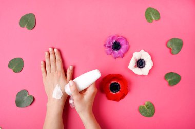 Çiçek ve yapraklarla pembe arka planda kozmetik krem sürmüş bir kadın. Kaplıca, güzellik ve tedavi konsepti. Düz yatış, üst görünüm