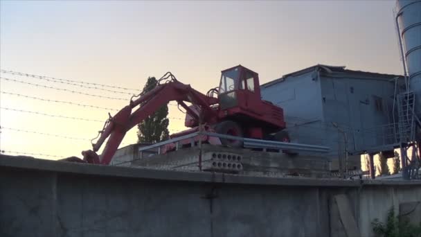 Sebuah ekskavator merah berdiri pada platform dekat pabrik. Penyimpanan mesin geraknya di belakang kawat berduri — Stok Video
