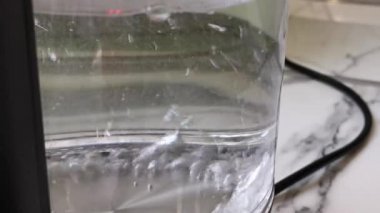 Su, elektrikli su ısıtıcısında kaynıyor. Mutfak masasında aydınlatma olan cam çaydanlık. Suyu kaynamaya getir. Su kabarcıklarının makro fotoğrafları. Doğal ışıkta kaynama süreci.