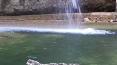 Su nehre yakın bir yere dökülüyor. Şelaleye ateş etmenin alt açısı ve oradan su sıçrıyor. Parlak turkuaz berrak dağ suyu. Taştan bir banka ve ormanda sığ bir nehir..