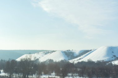 Winter landscape with field, trees and blue sky,long horizontal composition. Light fog and haze over the forest. Natural atmospheric cold background. View from the car window on the snow-covered trees