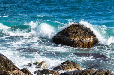 Turkuaz deniz taşı plajı, parlak güneş ışığında dalgalar kırılıyor. Renkli parlak deniz arka planı. Yaz konsepti, tatil, seyahat. Saf temiz deniz suyu, sahilde büyük taşlar.